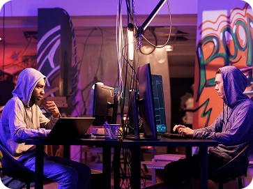 Deux personnes vêtues de sweats à capuche sont concentrées sur leurs ordinateurs dans une pièce faiblement éclairée, entourées de graffitis colorés, créant une atmosphère déterminée et intense.